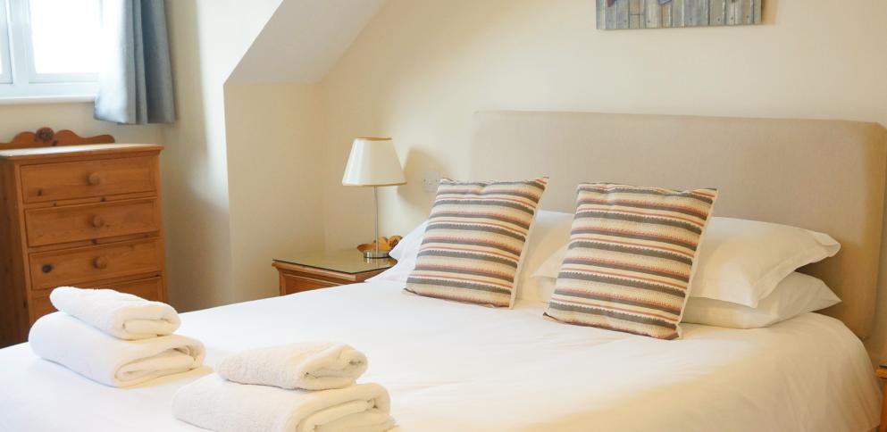 king bed with neutral headboard and two taupe/pink striped cushions.  Pine drawers in background